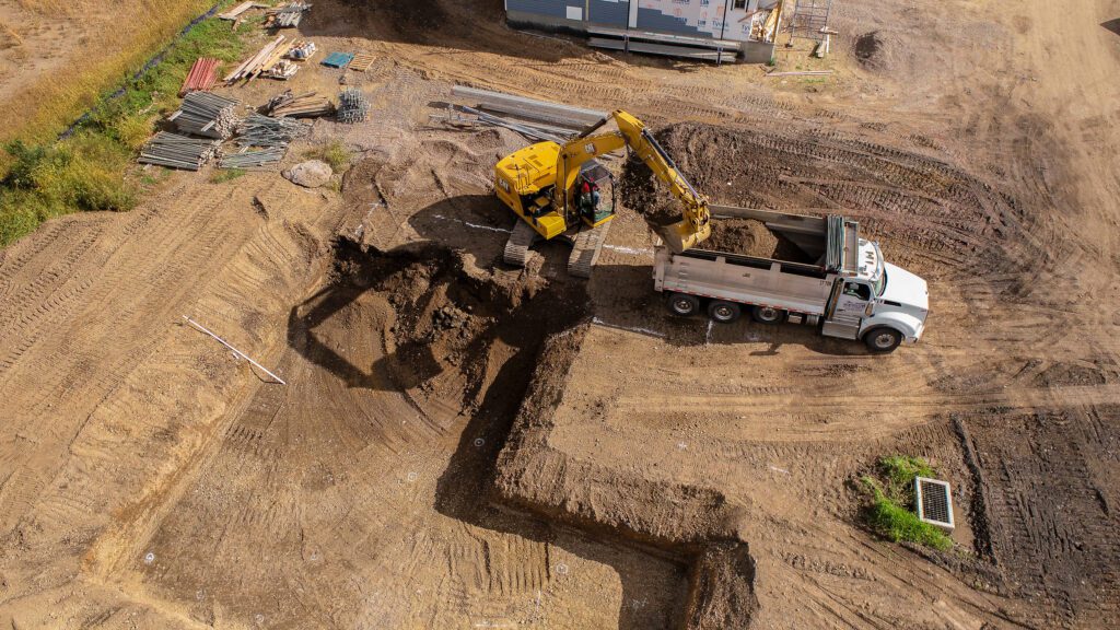 Residential construction company custom home construction shown from a drone view as an excavator and dump truck prepare the foundation, highlighting planning, precision, and expert site work.